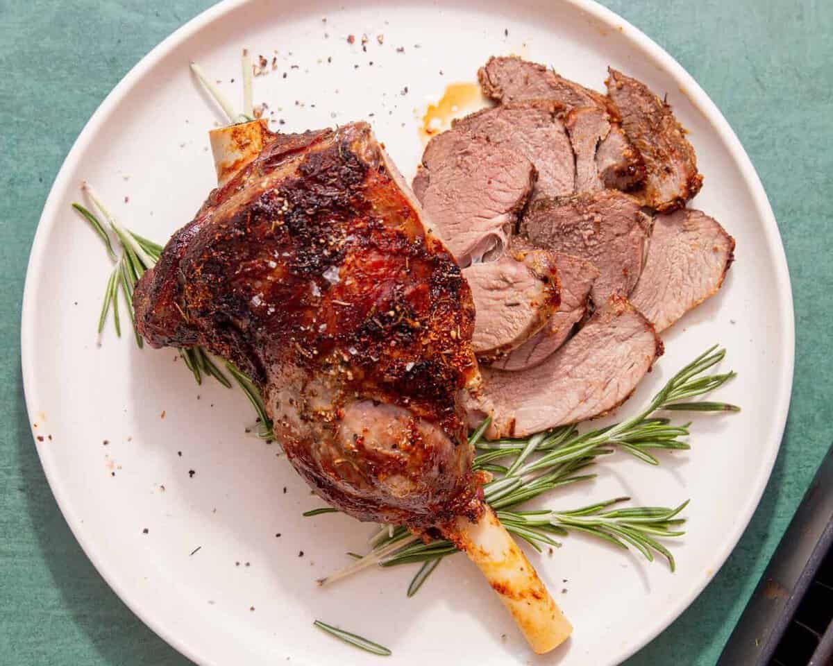 A cooked, seasoned leg of lamb is served on a white plate with a few slices cut, garnished with sprigs of rosemary, on a green surface.