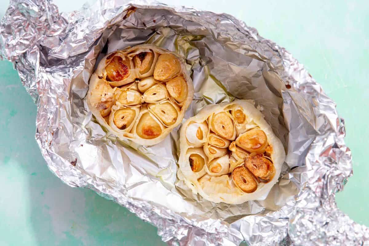 two roasted garlic bulbs in foil on a light green blue background