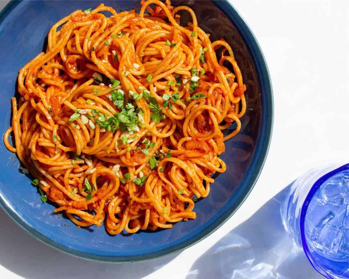 A blue bowl filled with tomatoey spaghetti topped with chopped parsley with blue glass filled with water/ice.