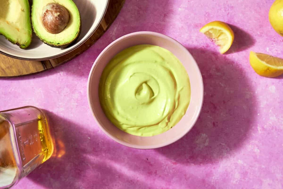 A bowl of creamy avocado sauce sits on a pink surface, surrounded by roasted corn, lemon wedges, a bottle, and a plate with halved avocados.