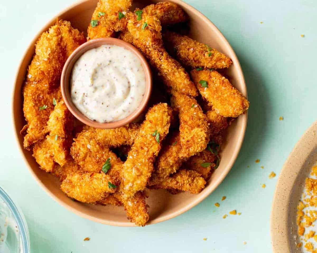 Overhead view of chicken goujons with dipping sauce on a plate, with partial view of cornflake bread crumbs on plate.