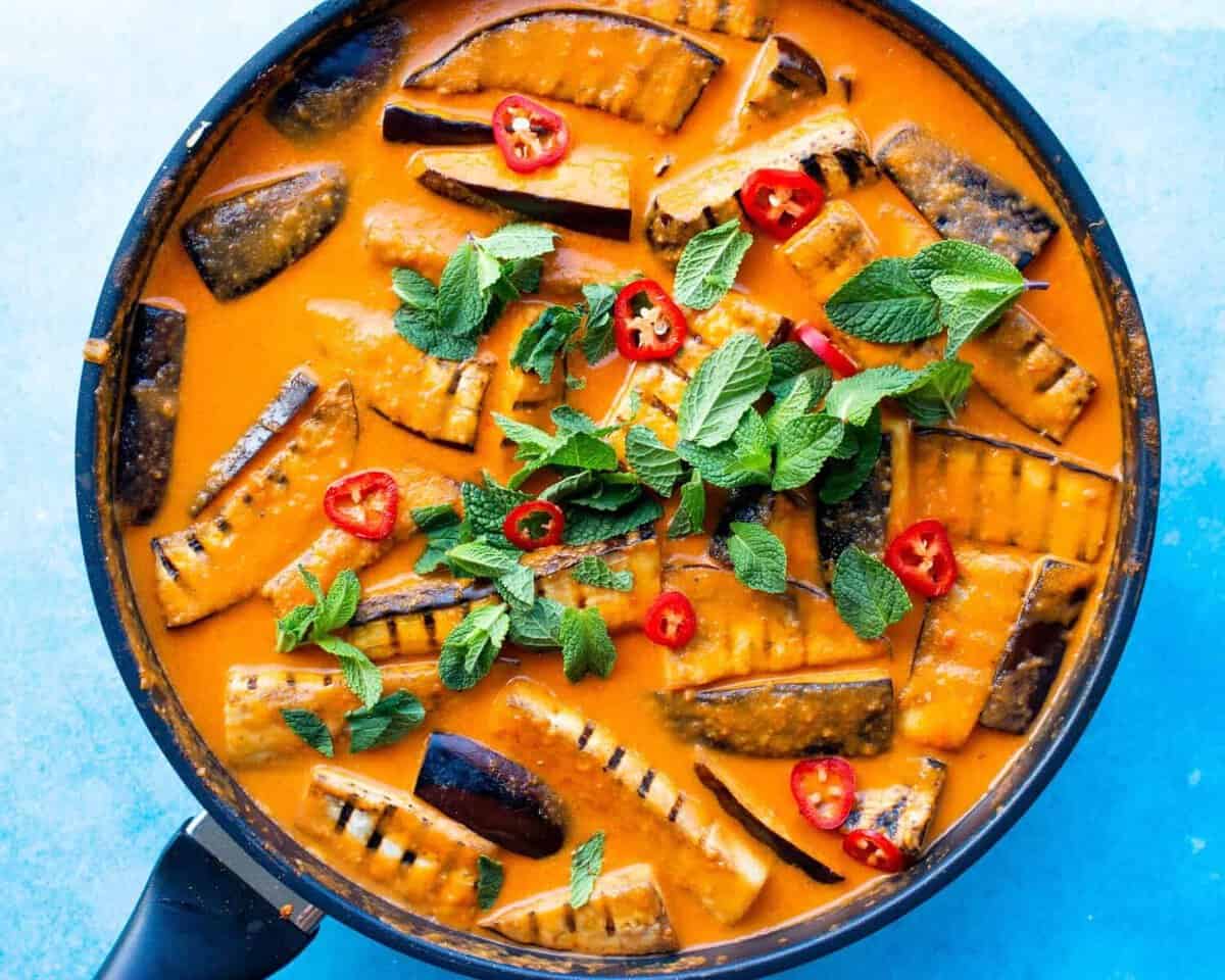 Aubergine curry in a large pan with aubergine charred batons covered in sauce topped with coriander and chillies.