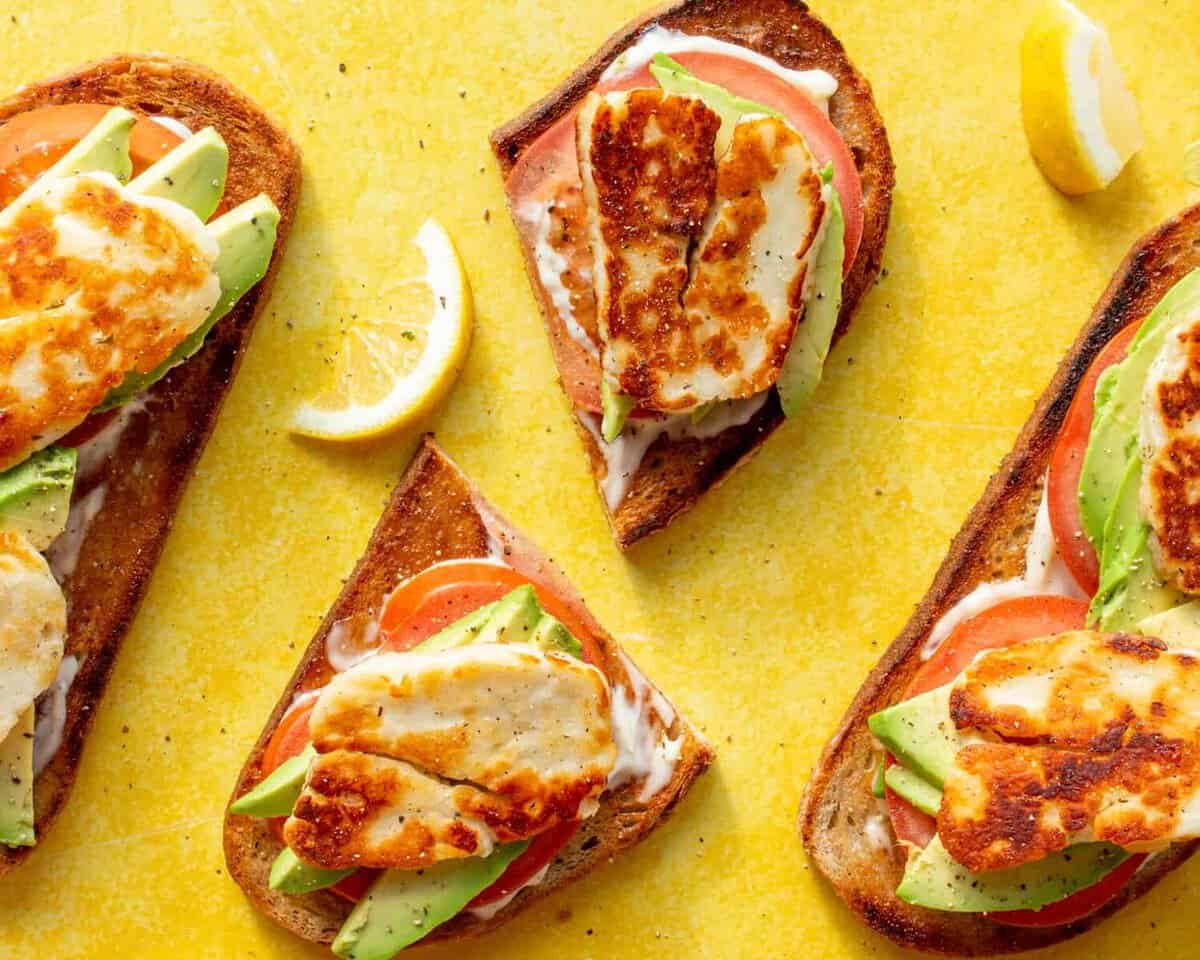 Halloumi and Avocado Sourdough Toast slices with avocado, tomato, mayonnaise topped with golden browned halloumi on a yellow background.