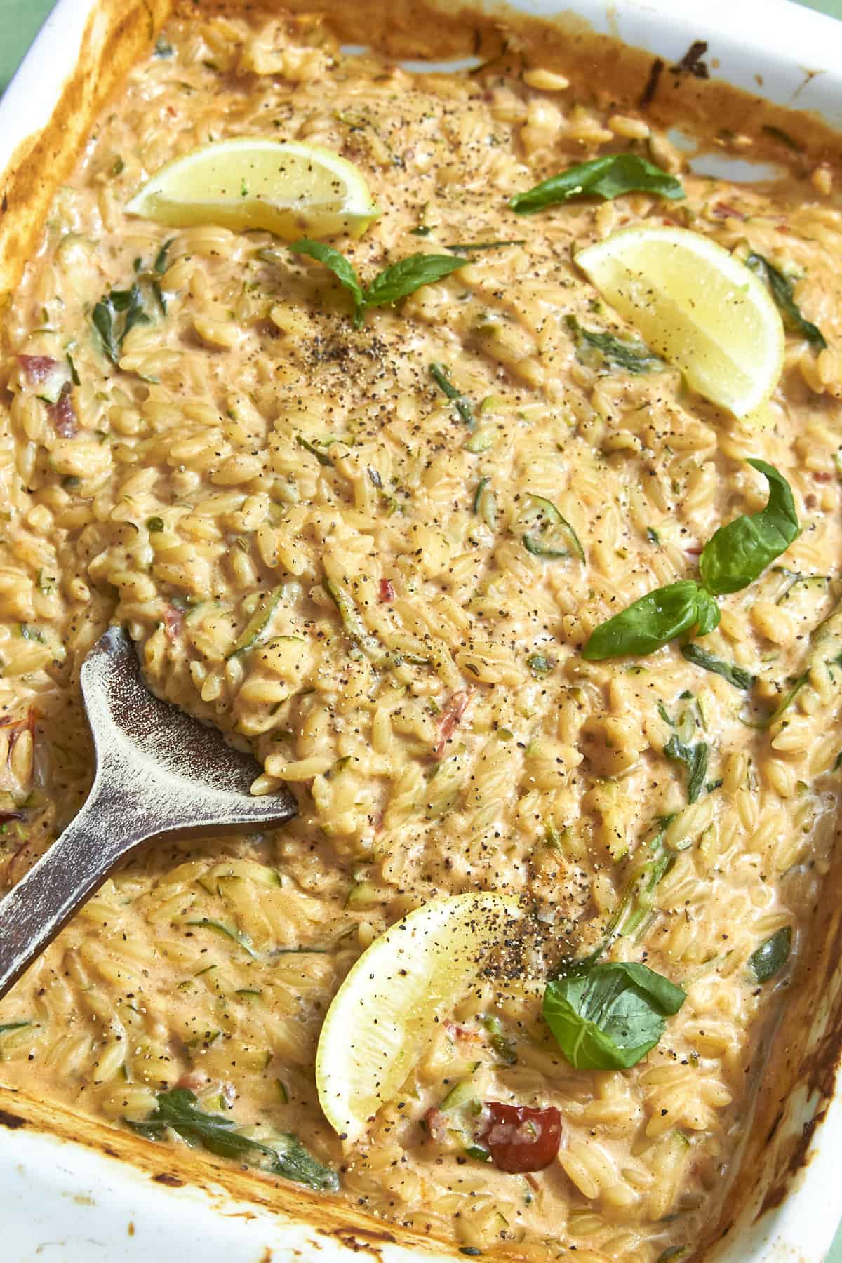 A creamy baked orzo dish with roasted corn, garnished with fresh basil leaves, lemon wedges, and cracked black pepper, with a serving spoon resting in the pan.