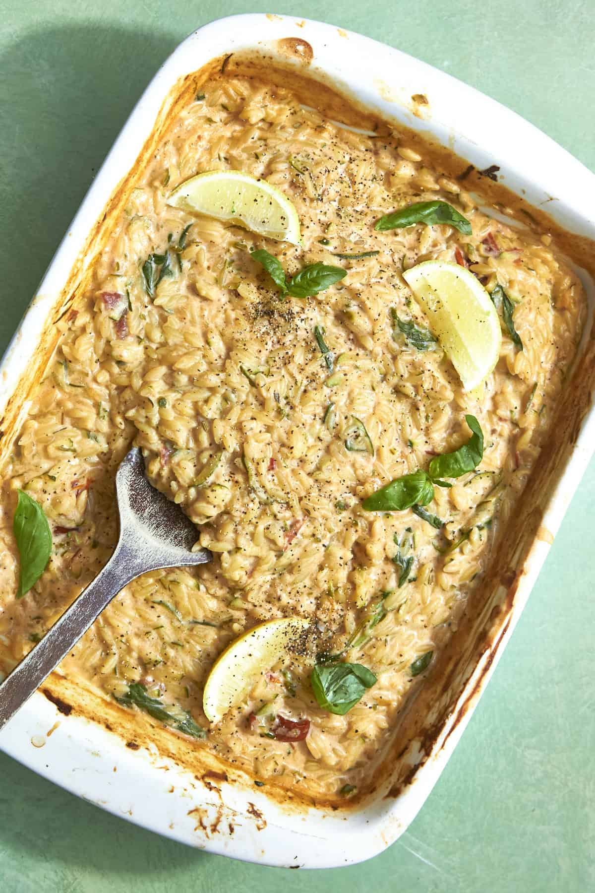 A baked creamy orzo pasta dish with roasted corn in a white rectangular pan, topped with lemon wedges, fresh basil leaves, and ground black pepper; a metal serving spoon rests in the pan.