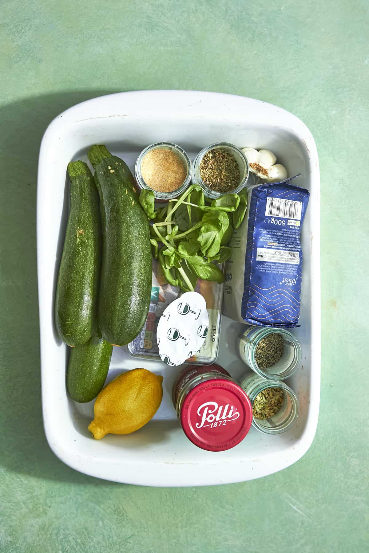 A white tray with two zucchinis, a lemon, basil, garlic, roasted corn, a jar of tomato sauce, a block of cheese, spices in small bowls, a bag of pasta, and dried herbs arranged on a light green surface.