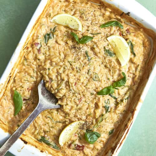 A white baking dish filled with creamy baked orzo and roasted corn, garnished with fresh basil leaves, lemon wedges, and black pepper. A serving spoon rests in the dish on a light green surface.