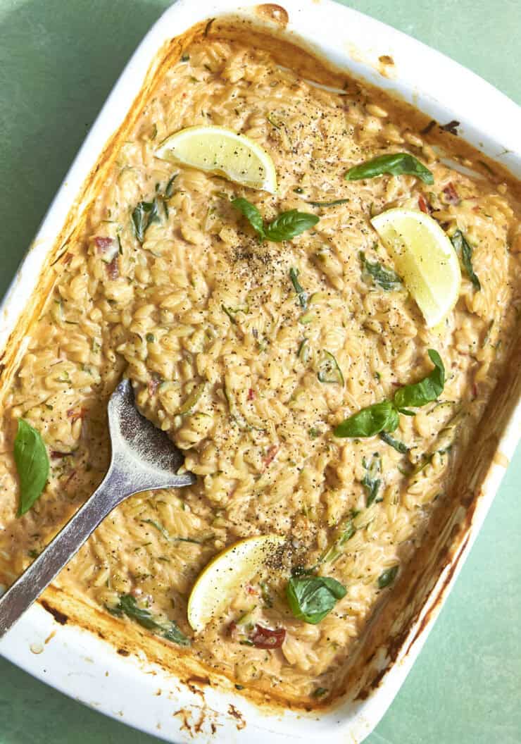 A white baking dish filled with creamy baked orzo and roasted corn, garnished with fresh basil leaves, lemon wedges, and black pepper. A serving spoon rests in the dish on a light green surface.