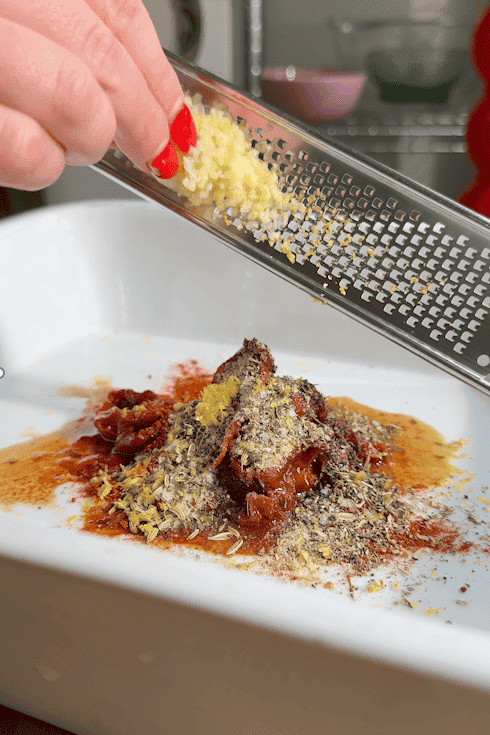 A hand grates fresh garlic with a microplane over a white dish containing herbs, spices, sun-dried tomatoes, and roasted corn, preparing ingredients for a recipe in a kitchen setting.