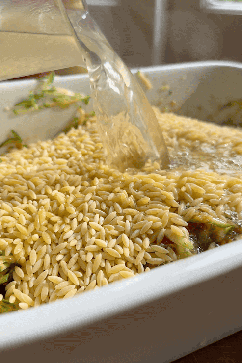 Close-up of uncooked orzo pasta and roasted corn in a white baking dish, with broth being poured over. Shredded vegetables are visible beneath the pasta, while steam rises from the hot liquid.