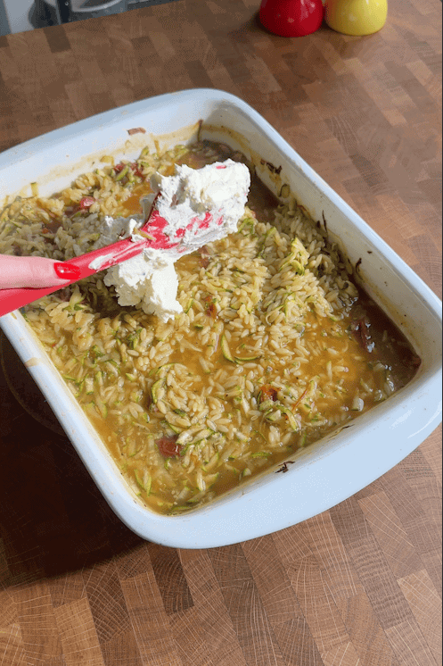A hand holding a red spatula with a scoop of ricotta cheese above a white baking dish filled with baked rice, roasted corn, and vegetables on a wooden countertop.