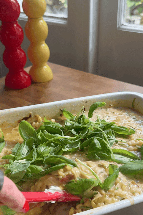 A hand stirs a baked pasta dish with roasted corn and fresh basil in a white baking tray on a wooden countertop. Red and yellow pepper grinders are in the background near a window.