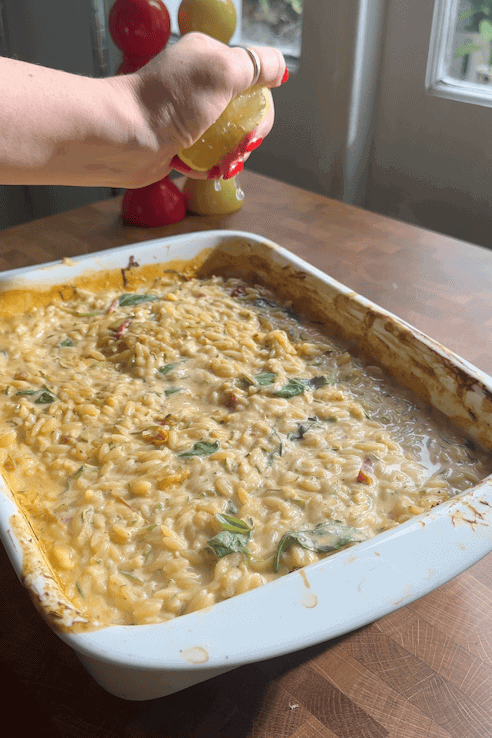 A hand squeezes a lemon over a baked orzo dish with spinach, roasted corn, and vegetables in a white casserole dish on a wooden countertop, next to red and yellow pepper grinders.