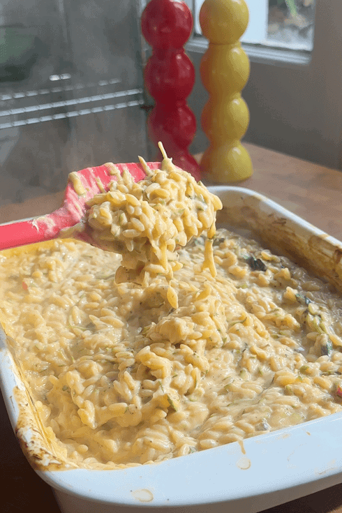 A close-up of a cheesy pasta bake with roasted corn being scooped from a white baking dish with a pink spatula, in a kitchen setting with red and yellow pepper grinders in the background.