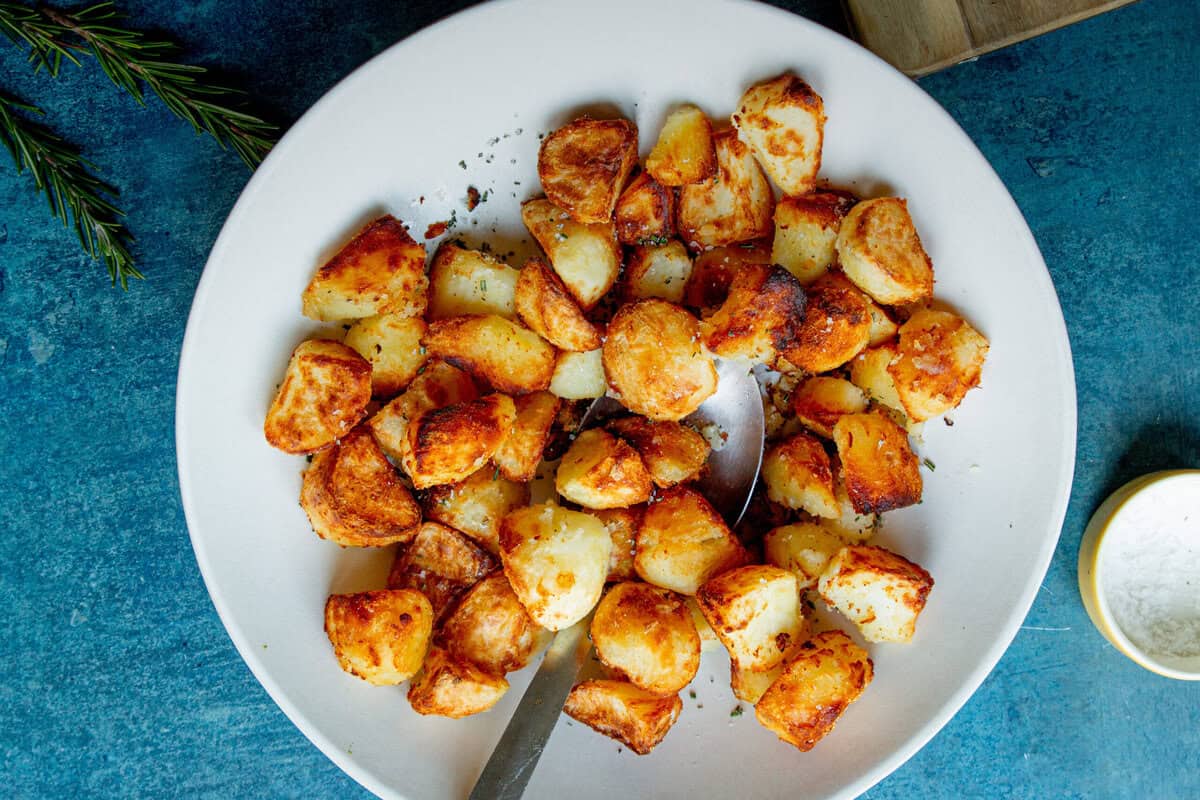 A white plate filled with golden, crispy roasted potatoes sits on a blue surface, with a silver serving spoon. Rosemary sprigs and a small bowl of salt are nearby.