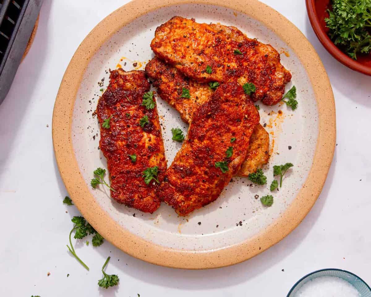 4 pieces of pork covered in a reddish sauce, some seasoning and some fresh parsley sprigs on a plate on a white background.