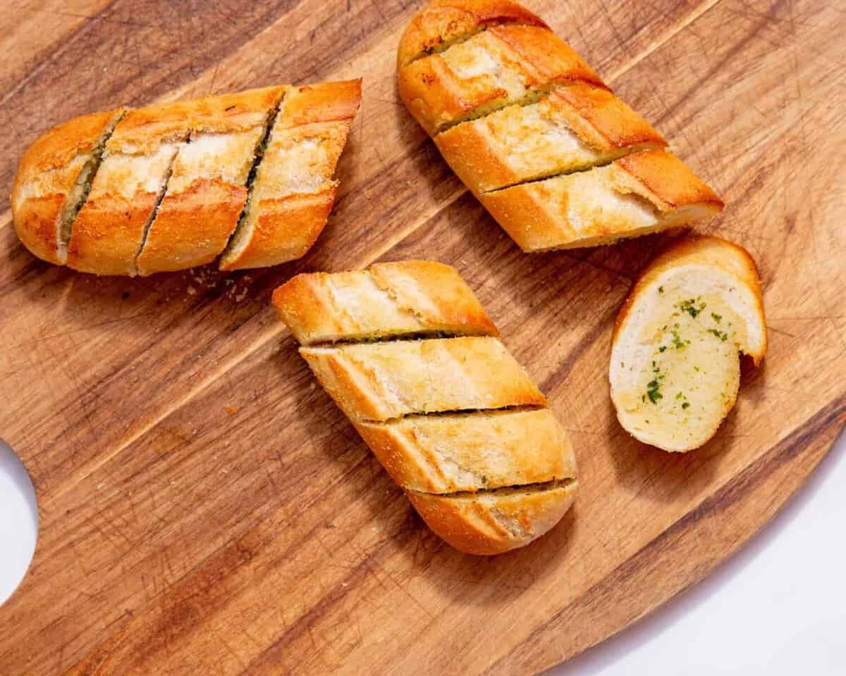 A golden browned, baked garlic bread baguette broken into 4 pieces with pre slice marks on a wooden chopping board.