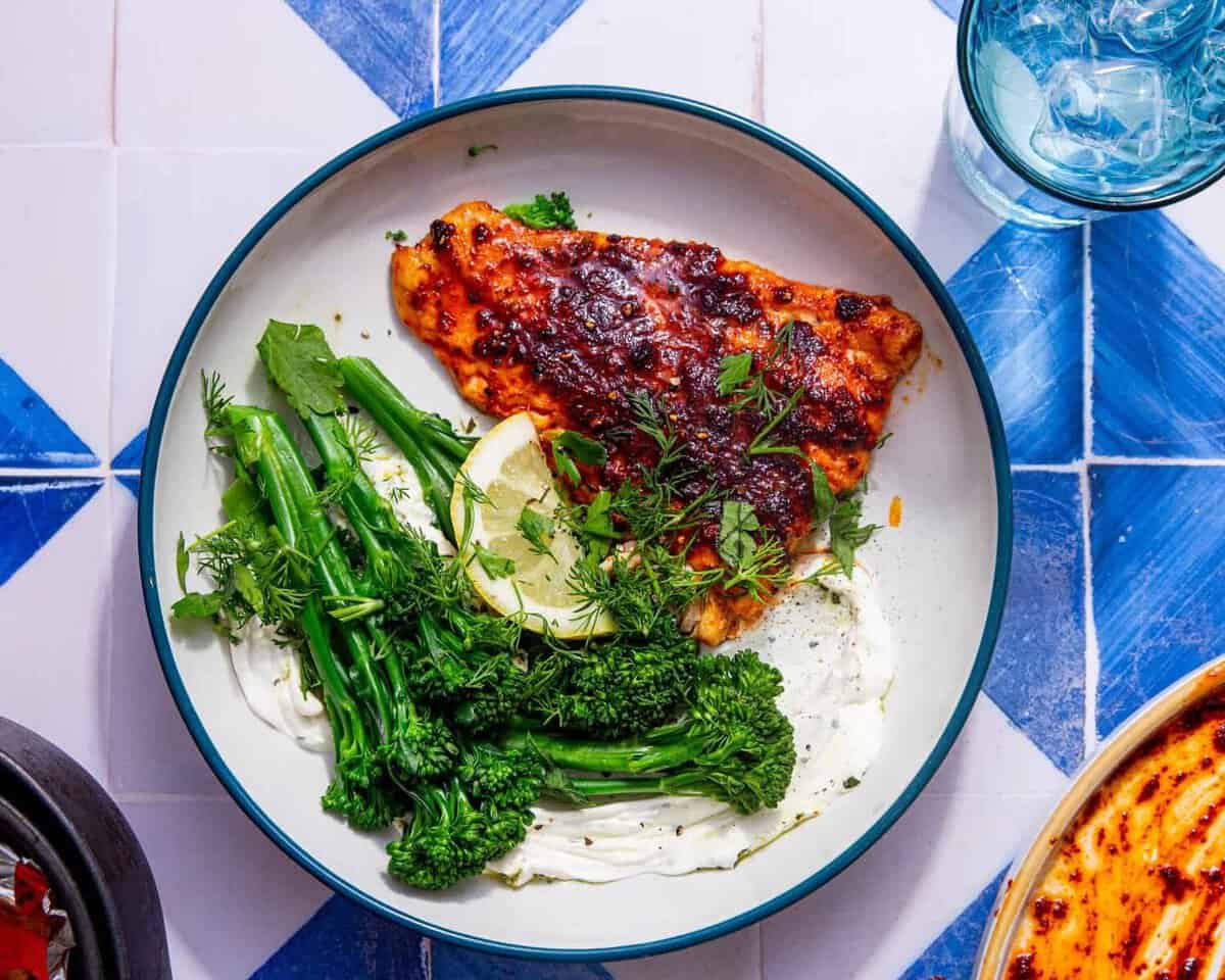 Well seasoned cod fillet on a white plate with tenderstem broccoli and a lemon wedge all on a blue and white tile background.