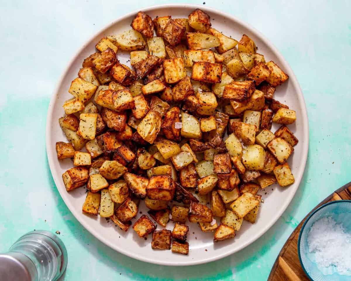 Golden browned diced potatoes with some salt sprinnkled over them on a plate on a pale blue background with a small bowl of salt on a wooden board and a pepper mill next to the plate.