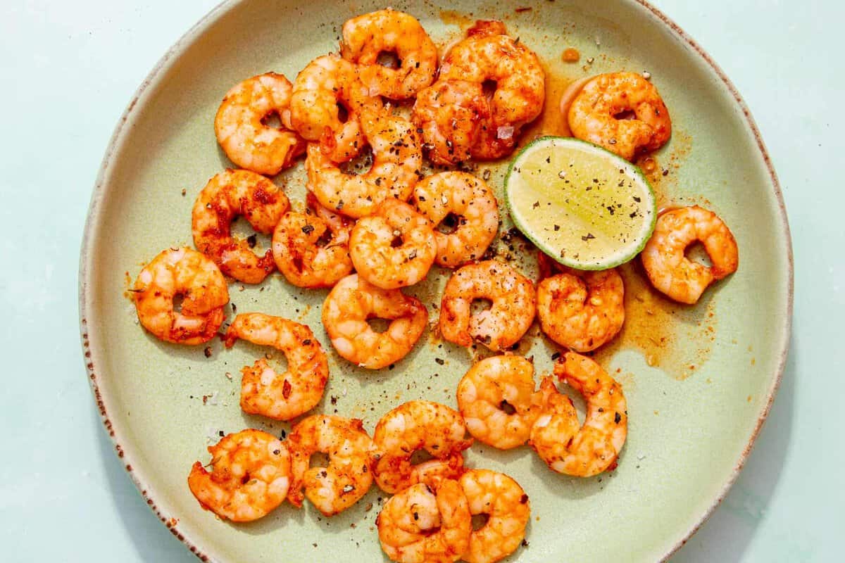 A beige plate with cooked, seasoned shrimp garnished with black pepper, served with a halved lime. The shrimp are arranged loosely, and the dish has a light, fresh presentation.