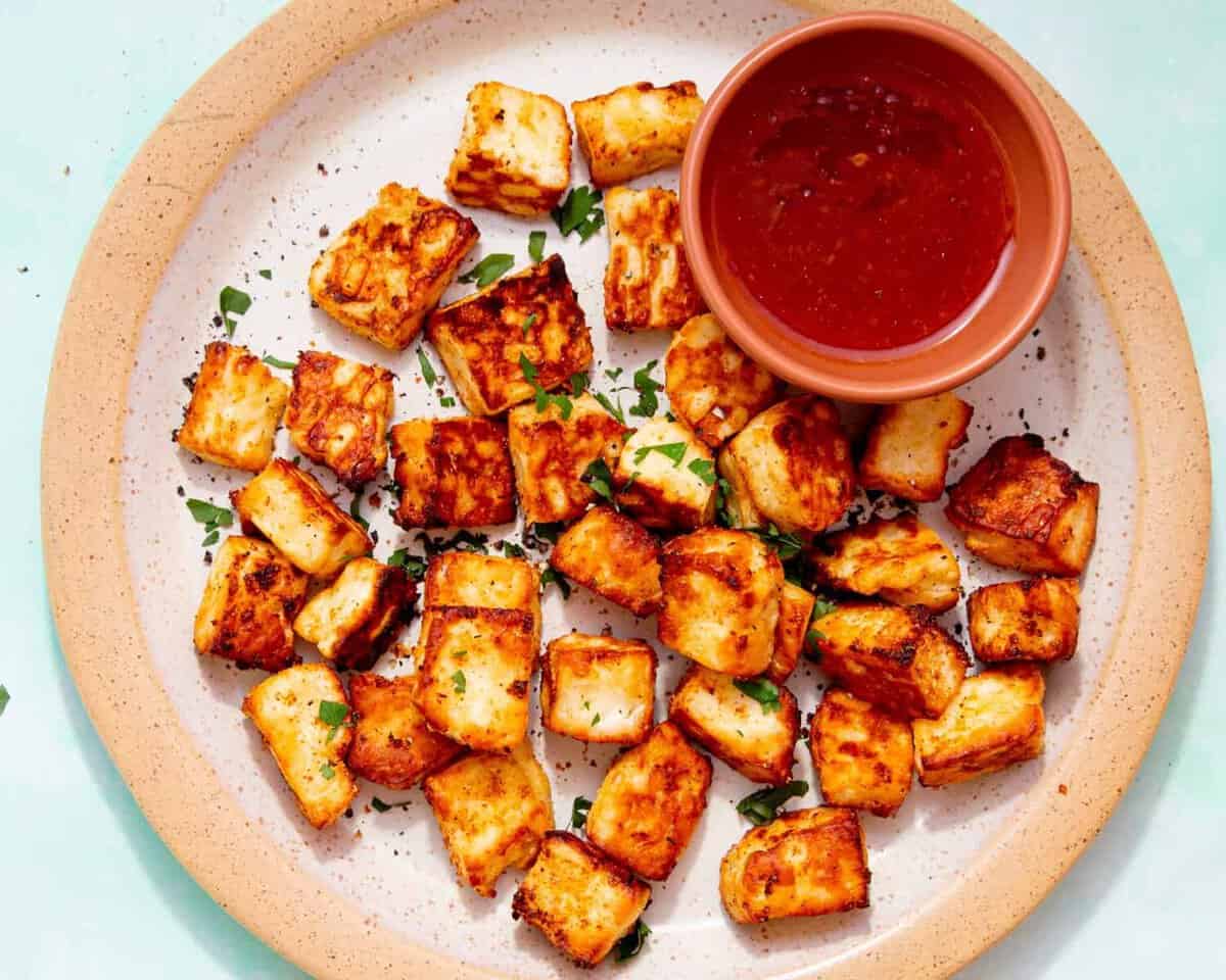 Golden browned halloumi cubes on a plate with some green herbs and a small bowl of chilli sauce on a pale blue background.