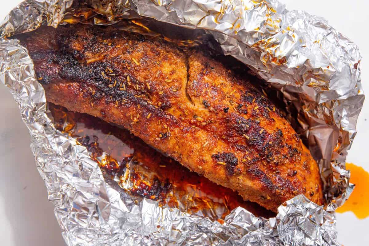 A cooked, seasoned pork tenderloin rests in crinkled aluminum foil, displaying a browned, flavorful crust with visible herbs and spices. Some orange-colored juices pool at the bottom of the foil.