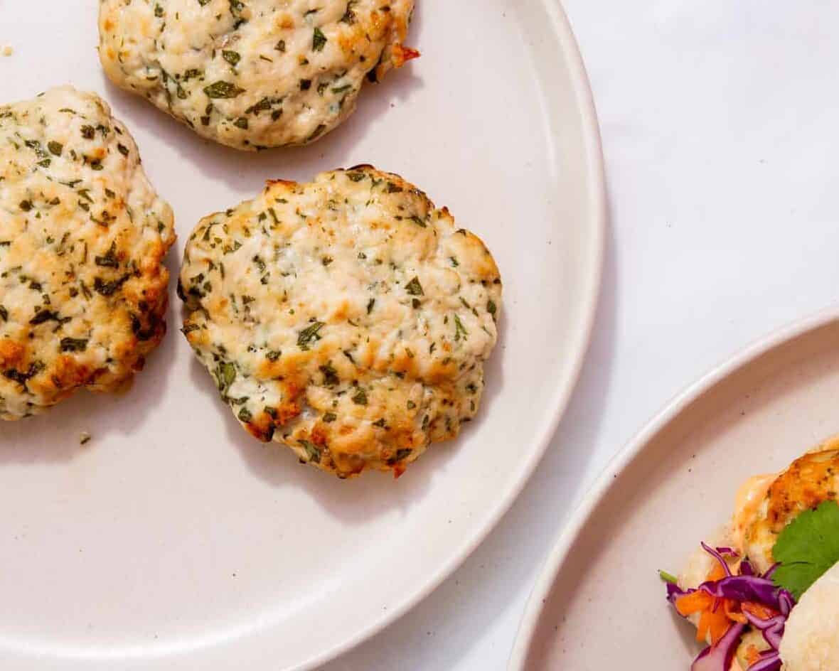 Three baked, golden-brown seafood cakes with herbs are arranged on a light-colored plate, with another plate partially visible in the corner, holding more seafood cake and a colorful edible flower garnish.