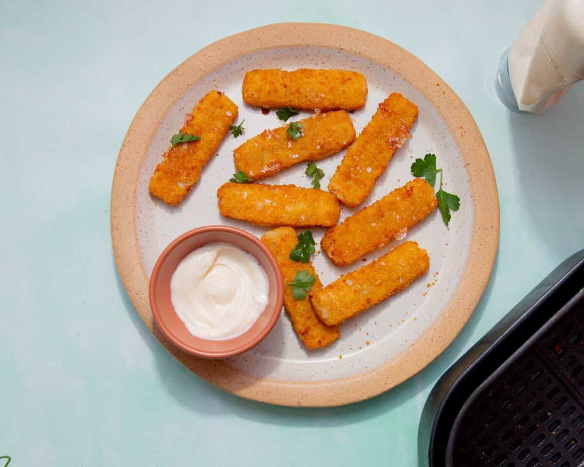 8 golden browned battered fish fingers on a plate with some mayonnaise in a small bowl and some fresh parsley.