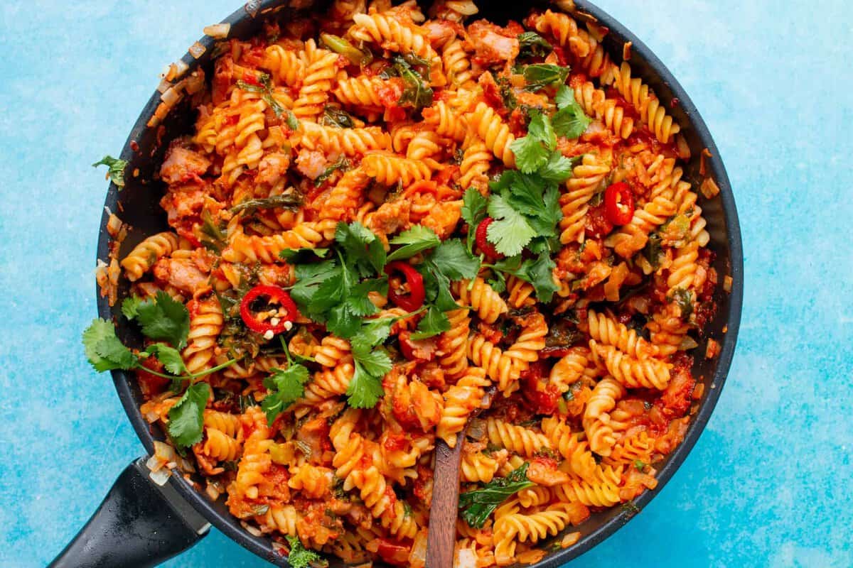 Fusilli Arrabiata with Veggie Sausages topped with red chillies and coriander in a large pan on a blue background.