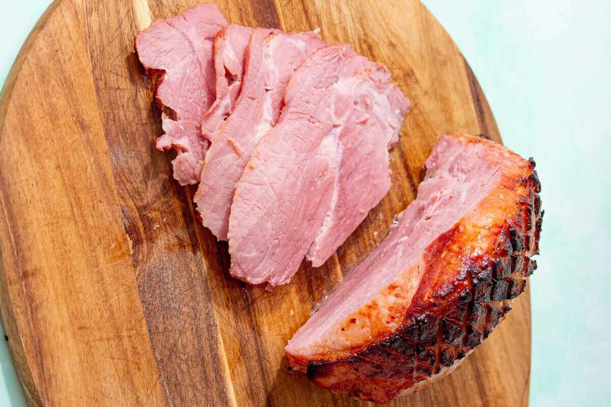 A partially sliced smoked ham sits on a wooden cutting board. Several thin slices are fanned out next to the unsliced portion, showing the juicy, pink interior and caramelized crust.
