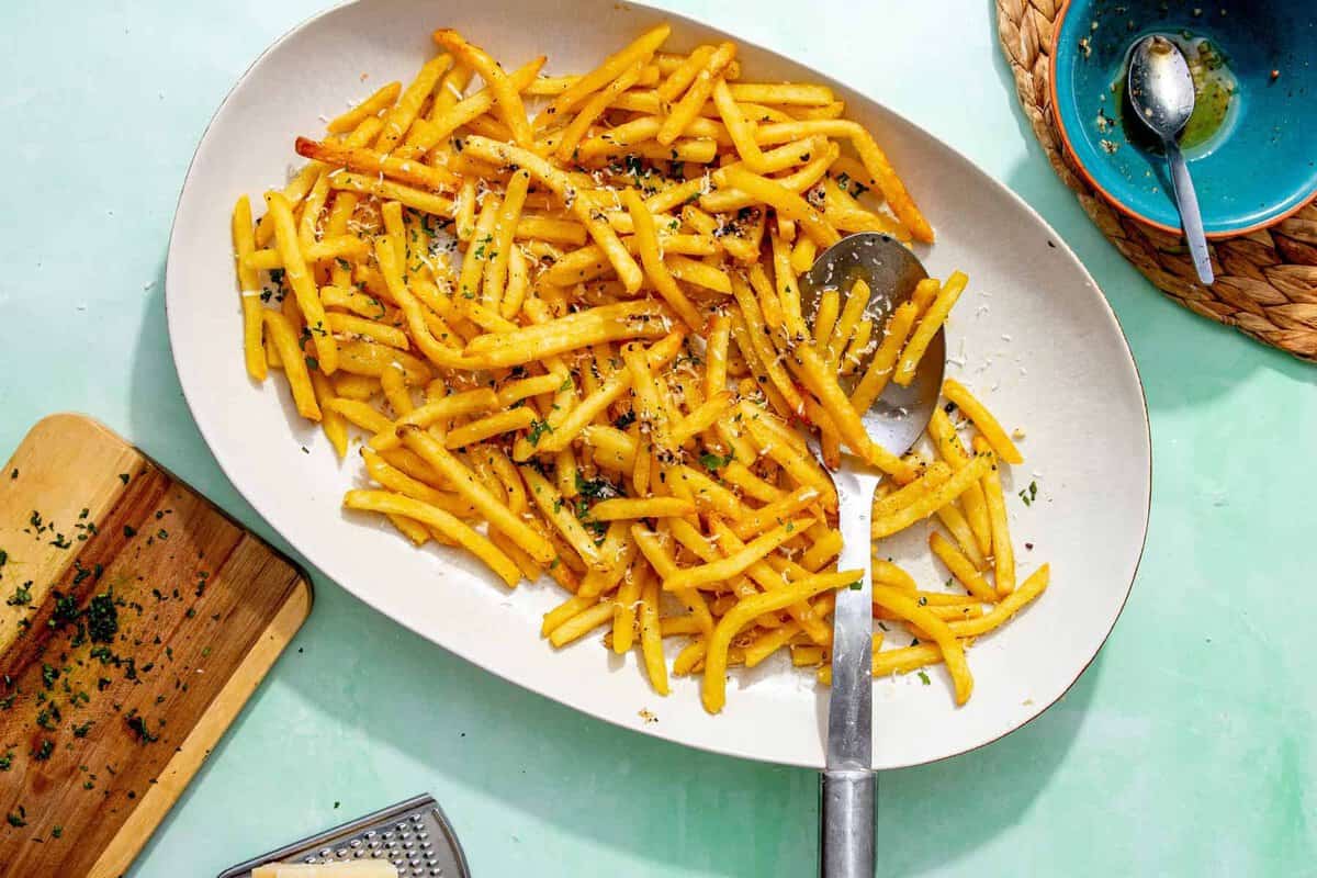 A platter of garlic fries topped with grated parmesan and chopped parsley, with a serving spoon beside them and a bowl of leftover garlic butter in the background.