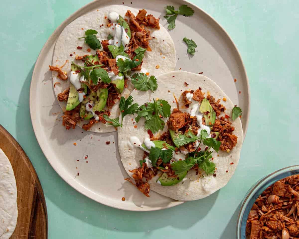 Three soft tacos on a beige plate, filled with shredded jackfruit, avocado slices, cilantro, a white sauce, and red pepper flakes. A bowl of jackfruit filling is partially visible nearby on a mint-green surface.