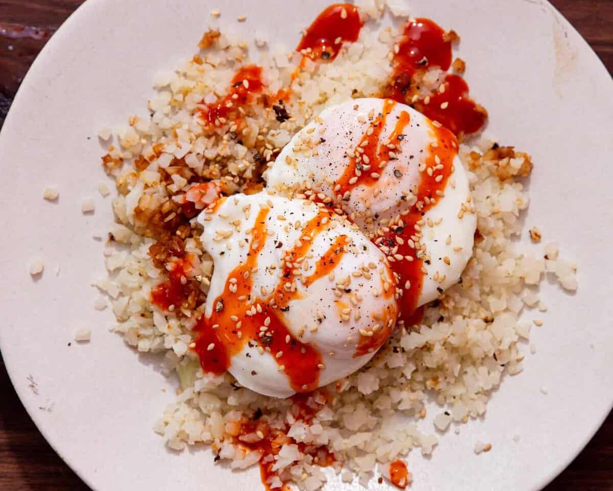 2 cooked poached eggs on a bed of rice with a drizzle of sriracha over the top on a white plate.