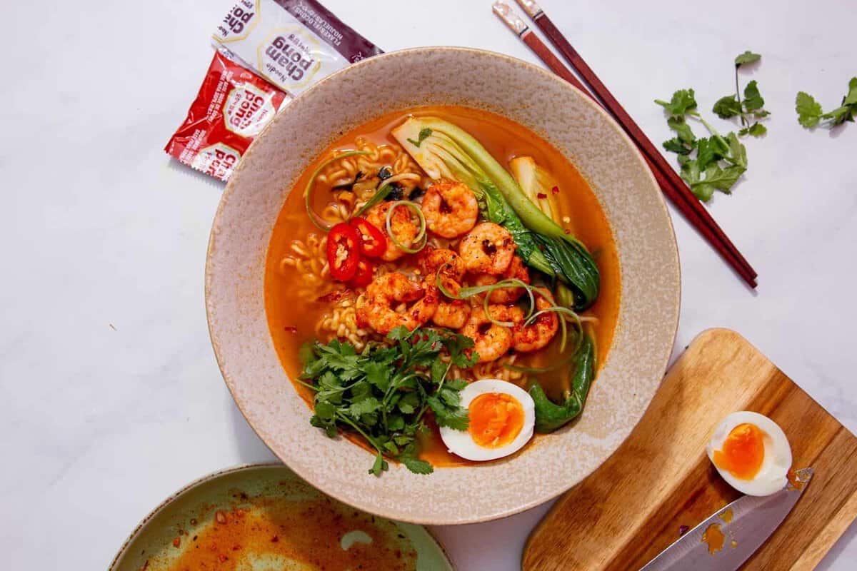 A bowl of shrimp ramen with bok choy, sliced red chili, green onions, cilantro, and a halved soft-boiled egg. Sauce packets, chopsticks, and herbs are on the side, with another egg half on a cutting board.