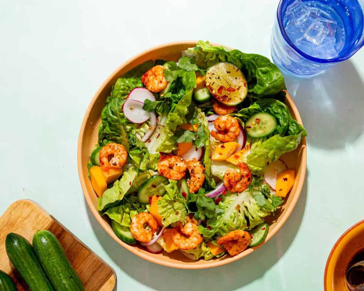 Golden browned king prawns in a bowl of salad with sliced cucumber, radished and red onion next to a blue glass with water.
