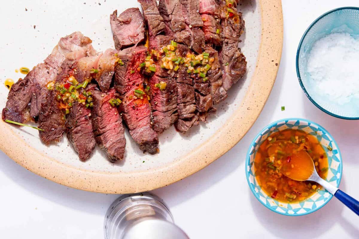 Sliced medium-rare steak on a plate, topped with a herb and chili sauce, with a bowl of dipping sauce and a bowl of coarse salt beside it.