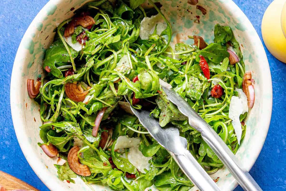 A bowl of fresh rocket salad with parmesan cheese, red onions, and sun-dried tomatoes, tossed with dressing. Metal tongs rest on the side of the bowl.