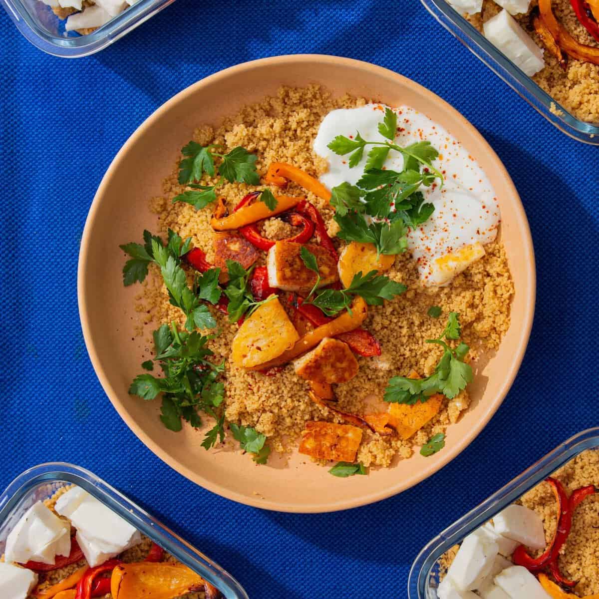 A bowl of spiced halloumi & couscous with peppers, yogurt and parsley and 4 meal prep containers with the halloumi, peppers and cous cous.
