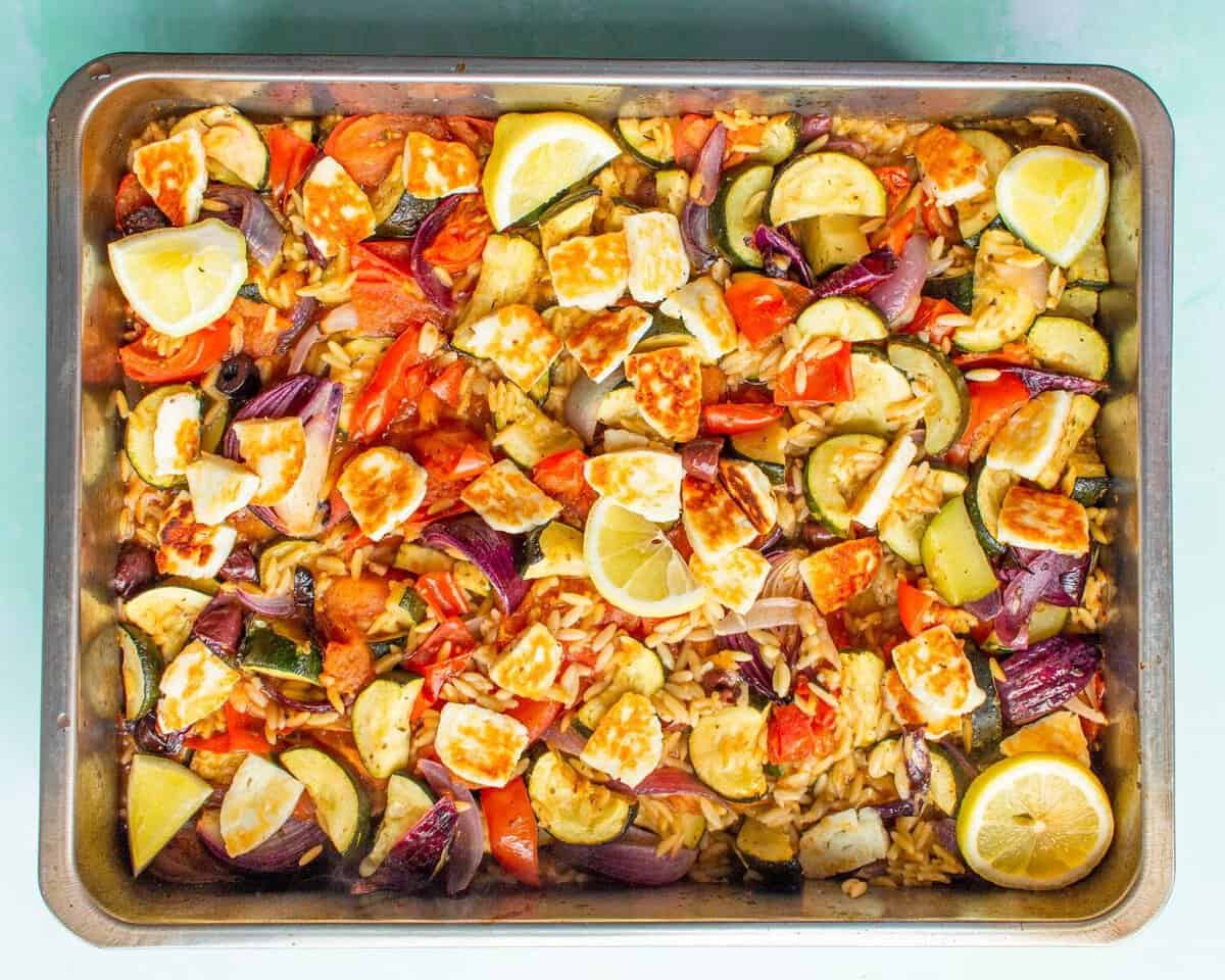 A baking tray filled with roasted vegetables including zucchini, tomatoes, red onions, and chunks of cheese, garnished with lemon wedges. The colorful dish sits on a light green surface.