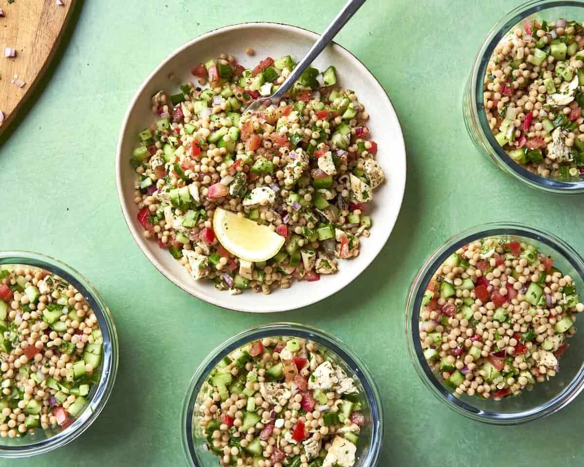 A plate and four bowls of couscous salad with diced cucumber, tomato, herbs, and feta cheese, garnished with a lemon wedge, are arranged on a green table. A fork rests on the main plate.