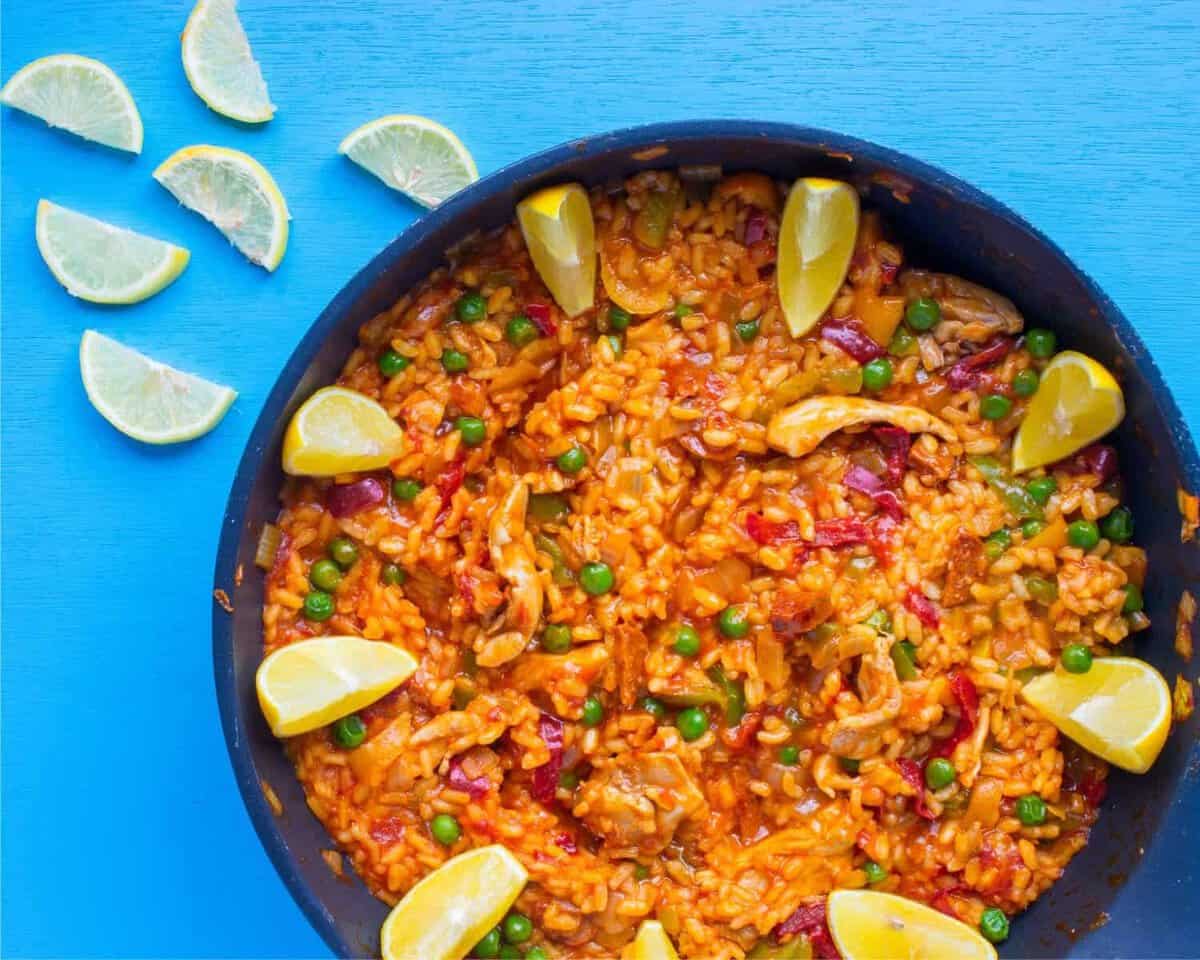 A skillet filled with yellow rice, peas, red peppers, and chunks of meat, garnished with lemon wedges, sits on a blue surface next to scattered lime slices.