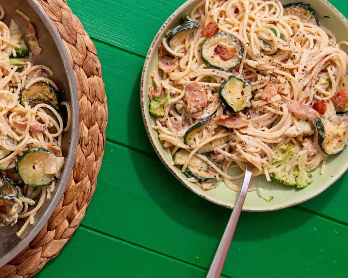 A bowl of creamy spaghetti, with bacon and courgettes with a spoon next to the spaghetti pan on a mat on a green background.