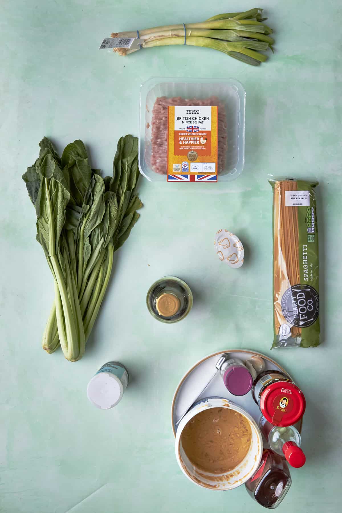 Ingredients for budget dan dan noodles laid out flat, including spaghetti, chicken mince, peanut butter and chilli oil.
