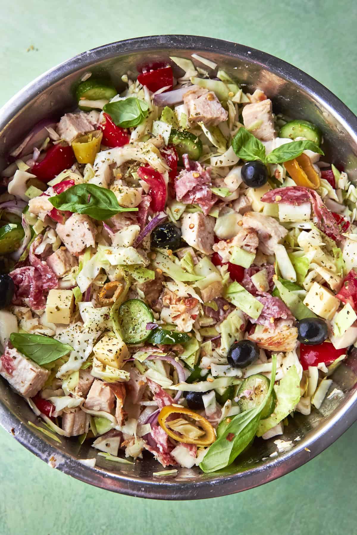 A bowl of Italian chopped salad with lettuce, cubed chicken, tomatoes, olives, cheese, basil, and sliced peppers, all mixed together on a green background.