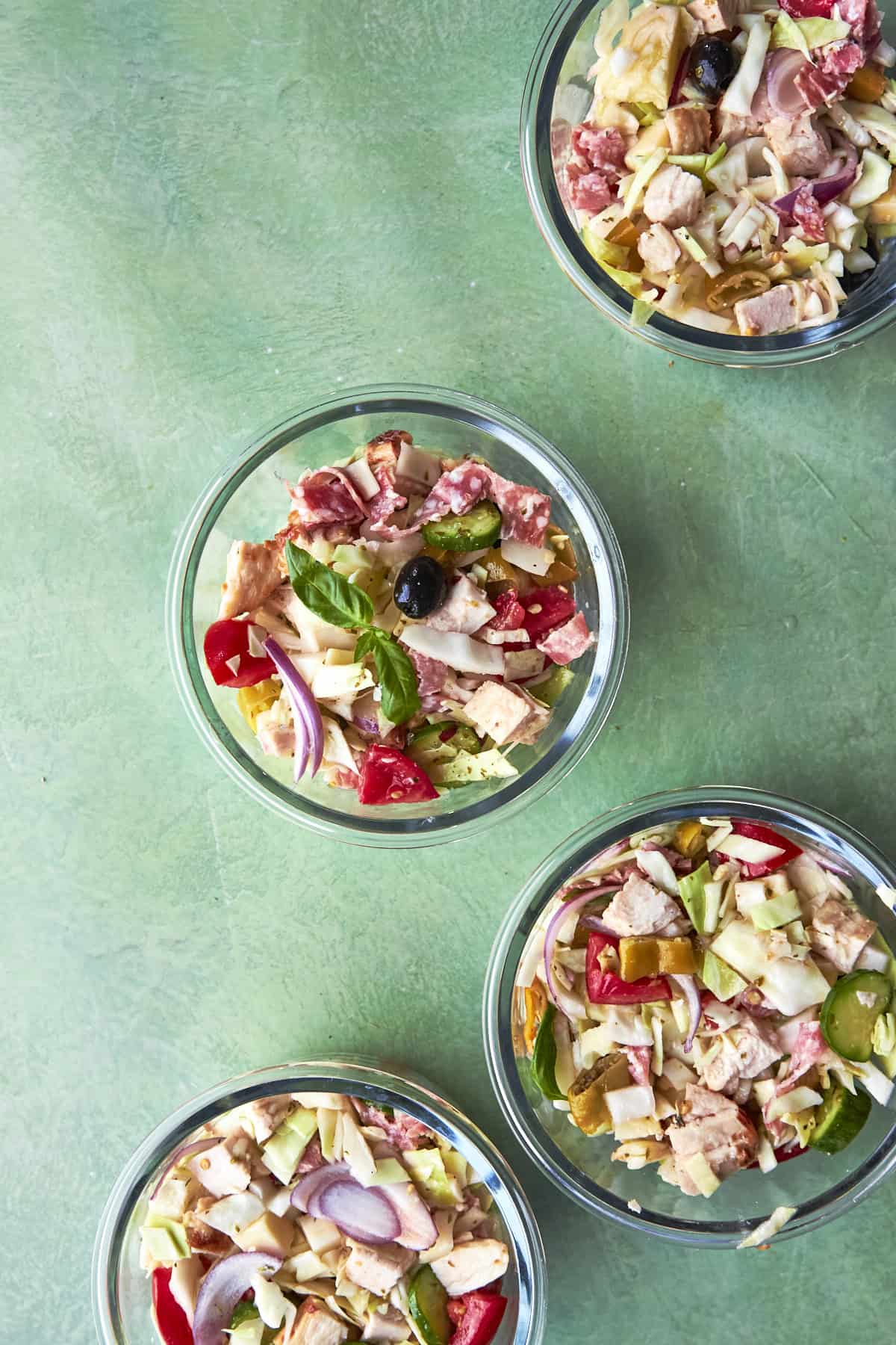 Four glass bowls filled with chopped salad ingredients like lettuce, tomato, olives, red onion, cheese, and deli meats, arranged on a green background.