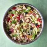 A metal bowl filled with chopped salad ingredients, including lettuce, tomatoes, olives, onions, cheese, tuna, and fresh basil, sits on a green surface.
