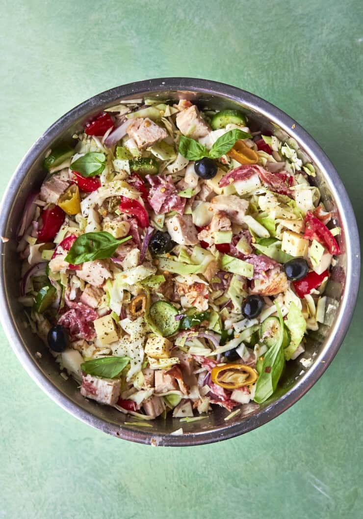 A metal bowl filled with chopped salad ingredients, including lettuce, tomatoes, olives, onions, cheese, tuna, and fresh basil, sits on a green surface.