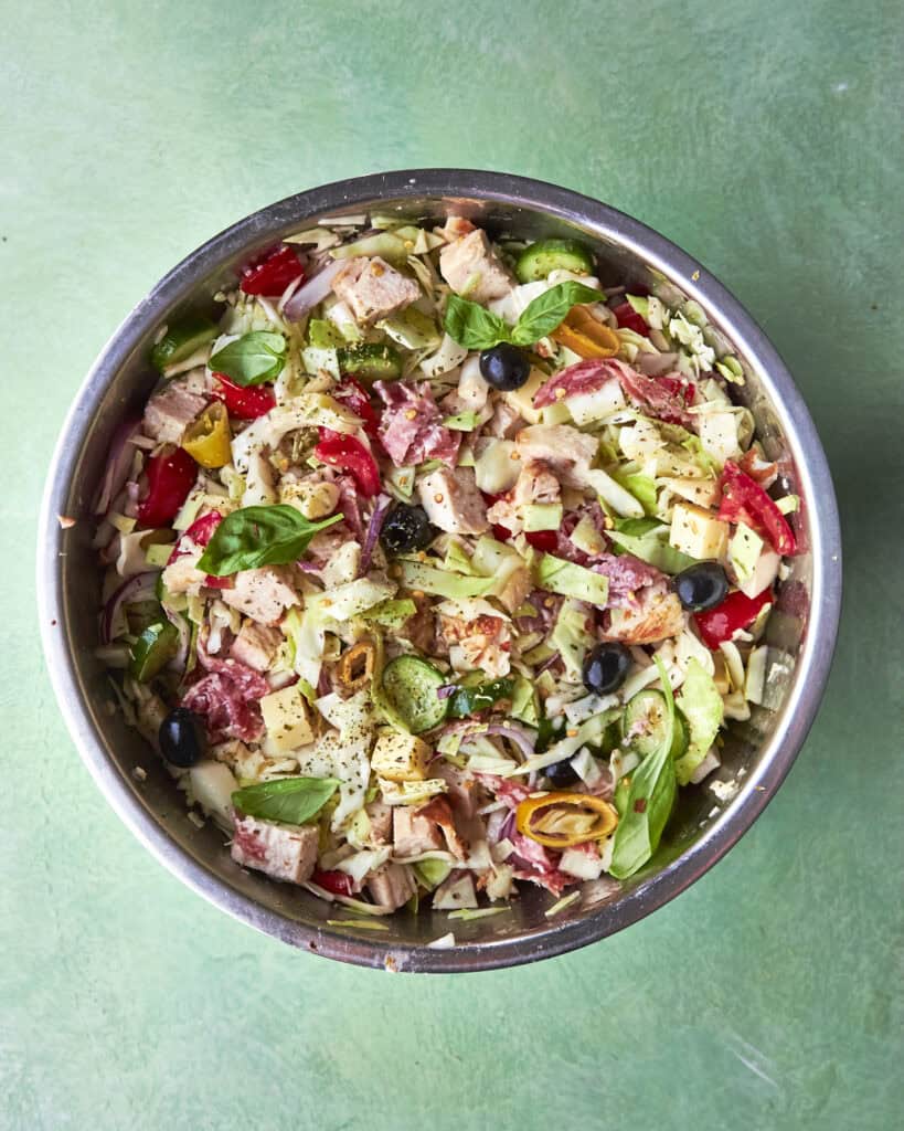 A metal bowl filled with chopped salad ingredients, including lettuce, tomatoes, olives, onions, cheese, tuna, and fresh basil, sits on a green surface.