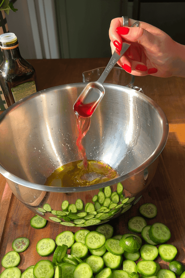 A hand pours red vinegar from a tablespoon into a metal bowl with oil. Sliced cucumbers are scattered on a wooden cutting board nearby. A bottle of olive oil is in the background.