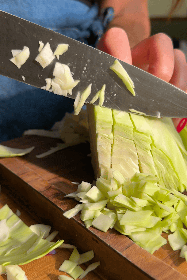 A close-up of a person slicing fresh green cabbage on a wooden cutting board with a sharp knife. The persons hand with red nail polish holds the cabbage in place.